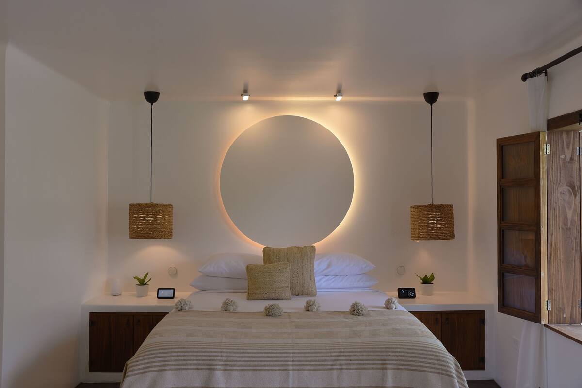 Ein Schlafzimmer mit einer runden Leuchte, Nachttischen mit Pflanzen, einem Bett mit gestreifter Bettwäsche und einem Holzschrank.