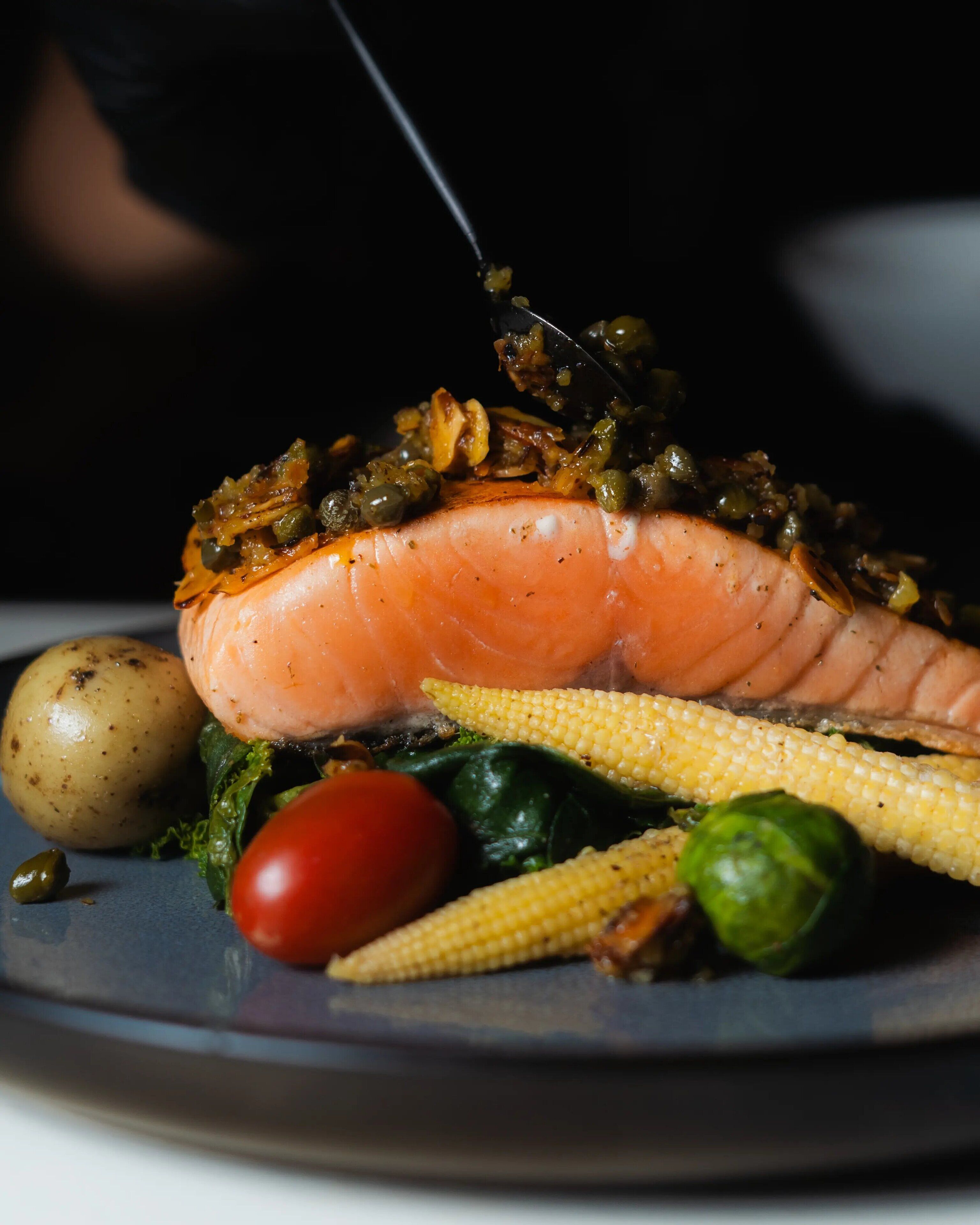 A plate with a salmon fillet, corn, a cherry tomato, a potato, and a green vegetable, likely spinach or lettuce.