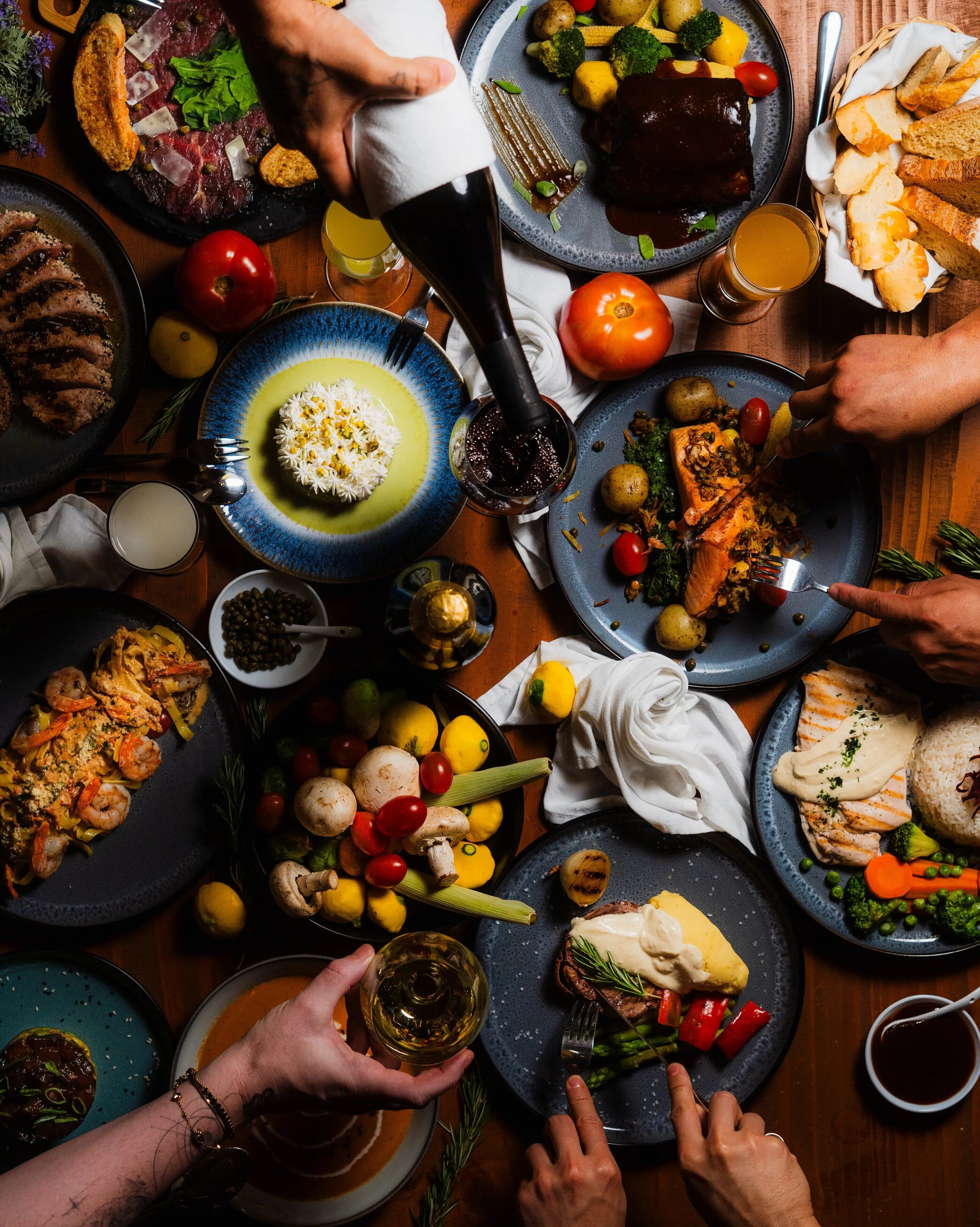 Variedad de platos en una mesa, incluyendo mariscos, verduras y carne, con gente comiendo y una botella de salsa siendo servida.