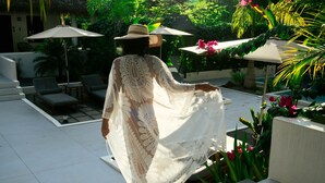 Une personne portant une robe à motifs se promène dans un espace extérieur ensoleillé, où se trouvent des chaises longues et des parasols.