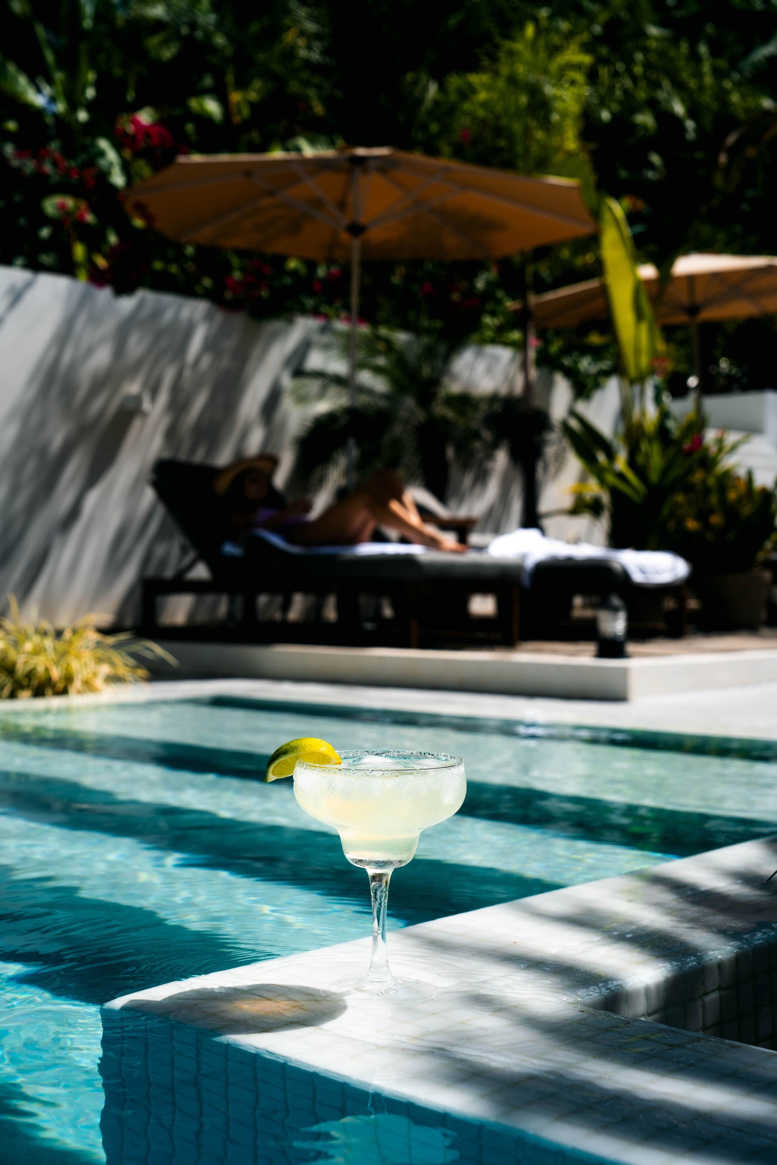Una piscina con un vaso de margarita en el borde, rodeada de sillas de descanso y sombrillas.