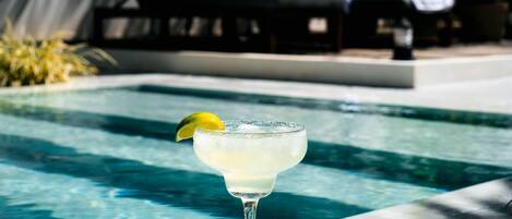 Una piscina con un vaso de margarita en el borde, rodeada de sillas de descanso y sombrillas.