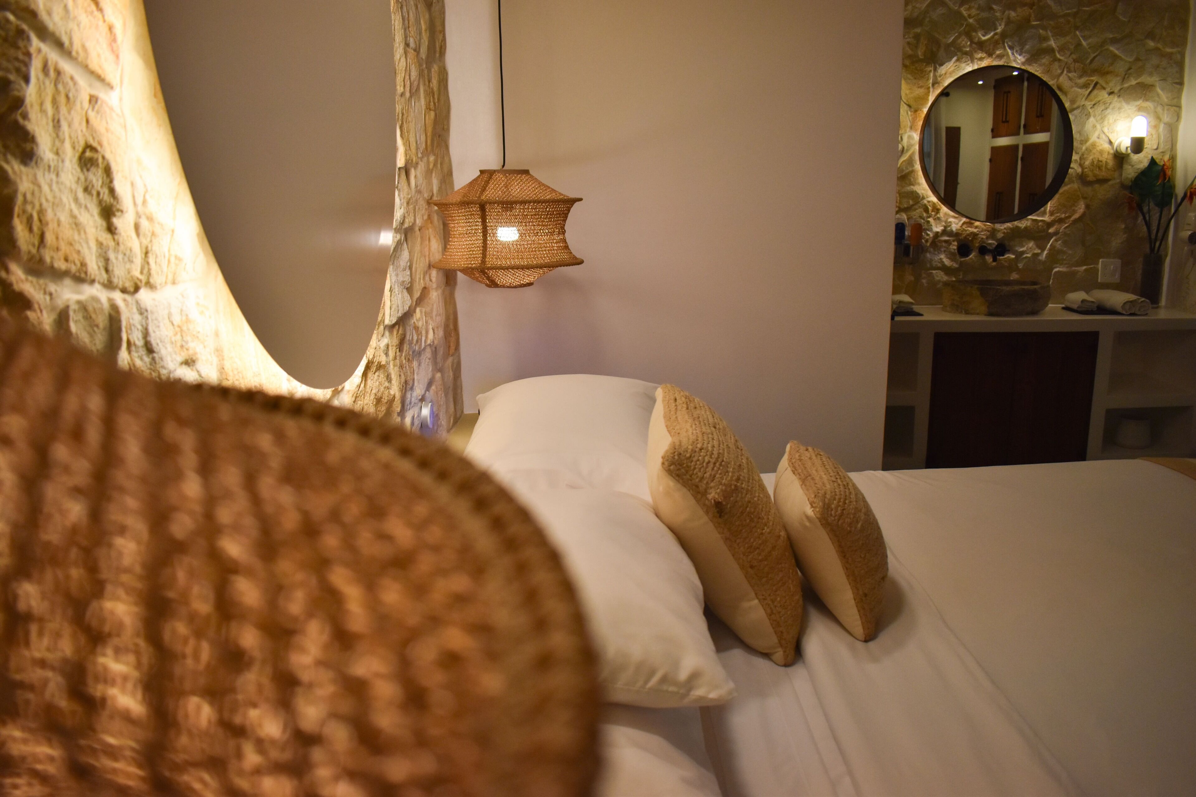 Ein Schlafzimmer mit Bett, Kissen, einer Hängelampe, einem runden Spiegel und einer Steinwand.