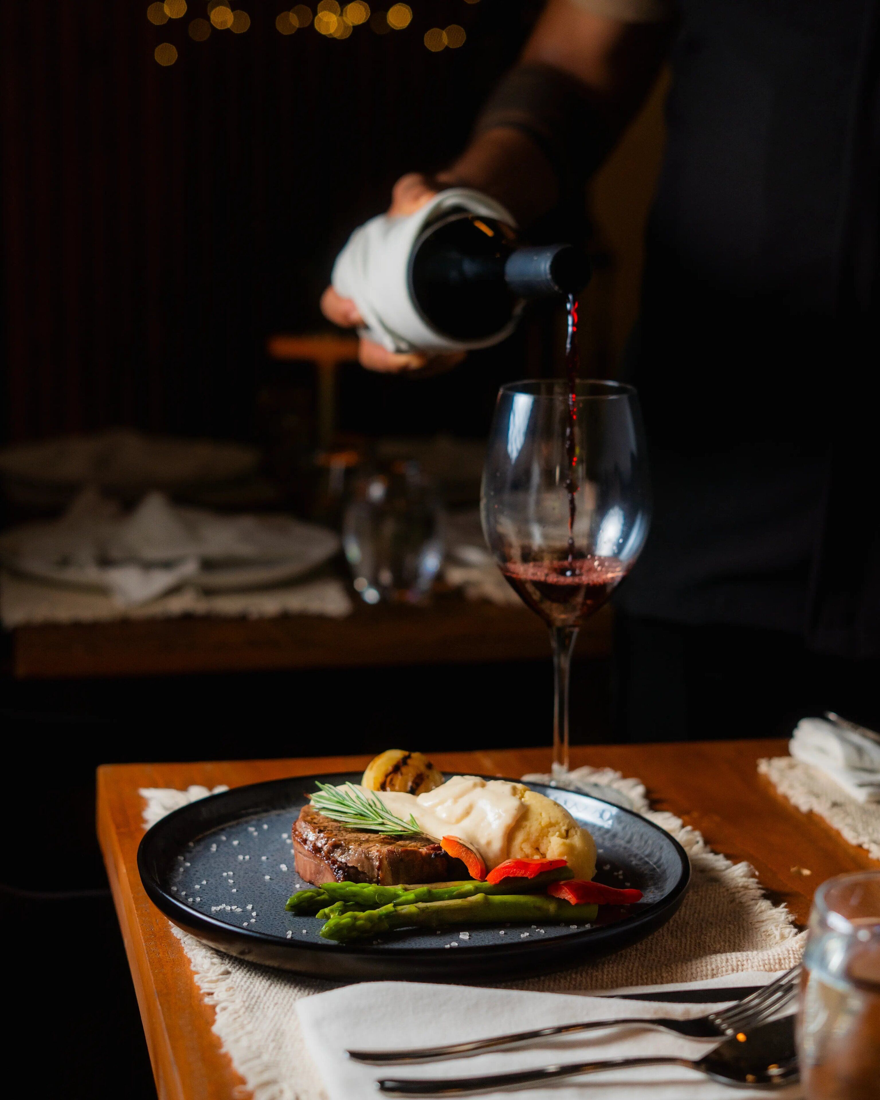 A dining table with a plate of food, a glass of red wine, and a person pouring wine.