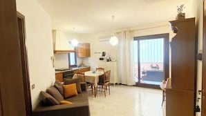 A living room with a sofa, dining table, and chairs. There is a kitchen area with cabinets and appliances. A sliding glass door leads to an outdoor area. - Two-room apartment in Olbia (Olbia)