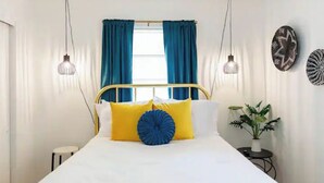 Ein Bett mit weißer Bettwäsche und zwei blauen Kissen, ein Nachttisch mit einer Pflanze sowie eine Wand mit zwei dekorativen Tellern.