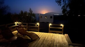 A wooden deck with a lounge chair, a small white building, and a view of the sunset over a mountain range. - Joshua Tree Hot Springs House (Twentynine Palms)