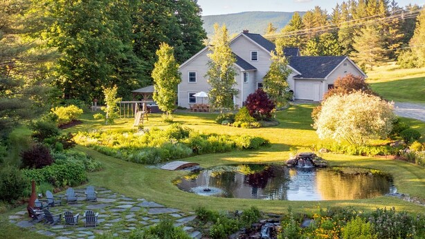 Ein großes Haus mit einem gepflegten Garten, einschließlich eines Teichs und einer Sitzgelegenheit im Freien.