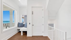 A hallway with a wooden floor, a white bench with blue cushions, a white door, and a white railing.