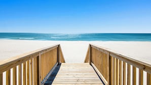 A wooden boardwalk leading to a beach with clear blue water and a bright sky.