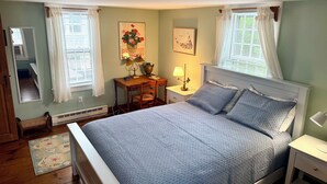 A bedroom with a bed, bedside tables, a nightstand, a mirror, a window with curtains, and a wall-mounted picture. - Charming Historic Downtown Cottage (Nantucket)