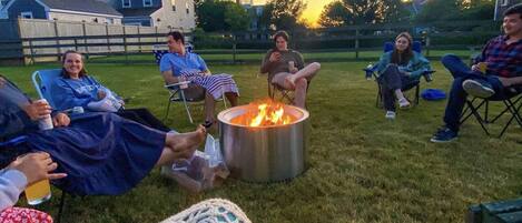 A group of people sitting around a fire pit in a grassy outdoor area with houses in the background.