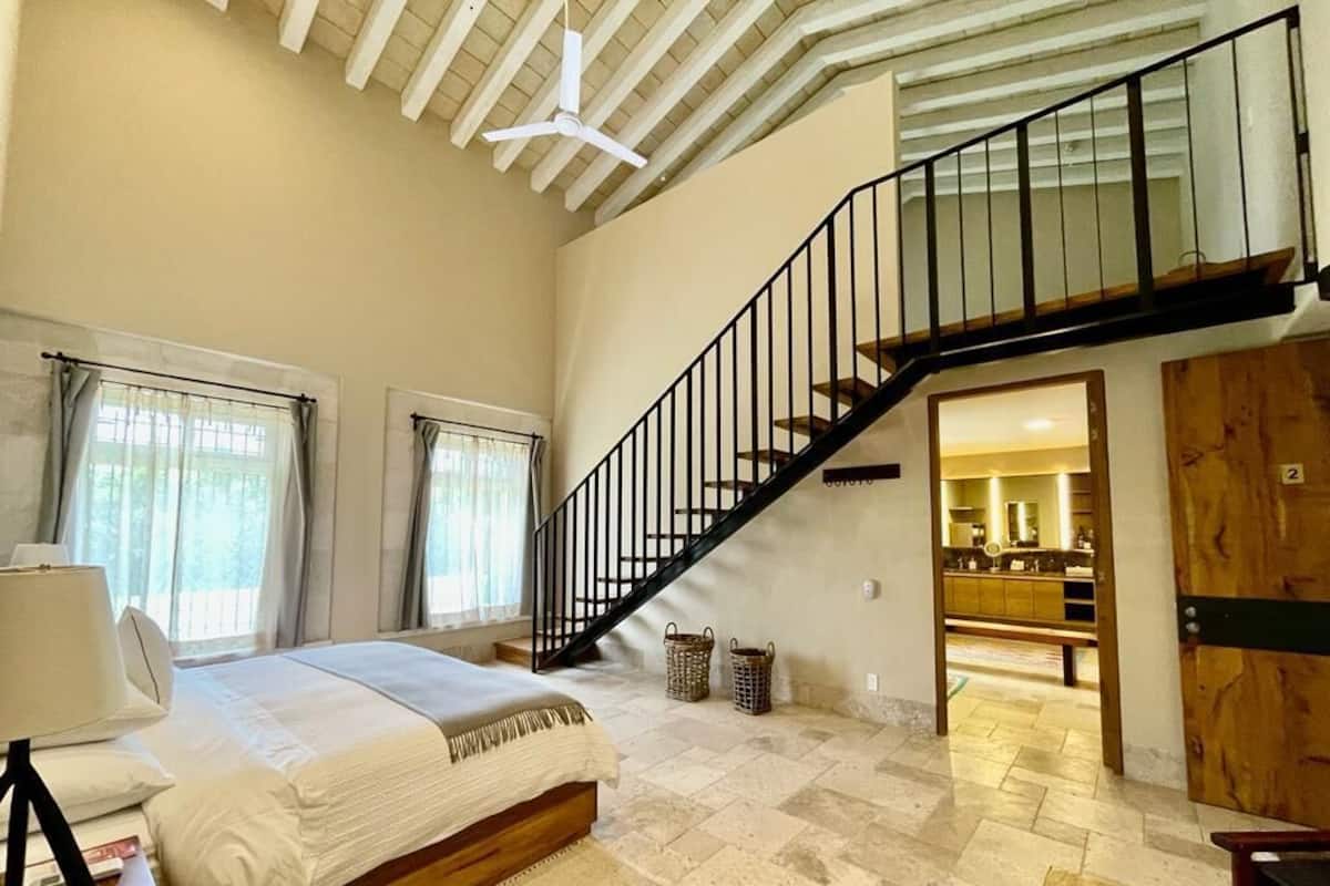 A spacious bedroom with a large bed, a wooden headboard, a staircase, and a view into the kitchen area.