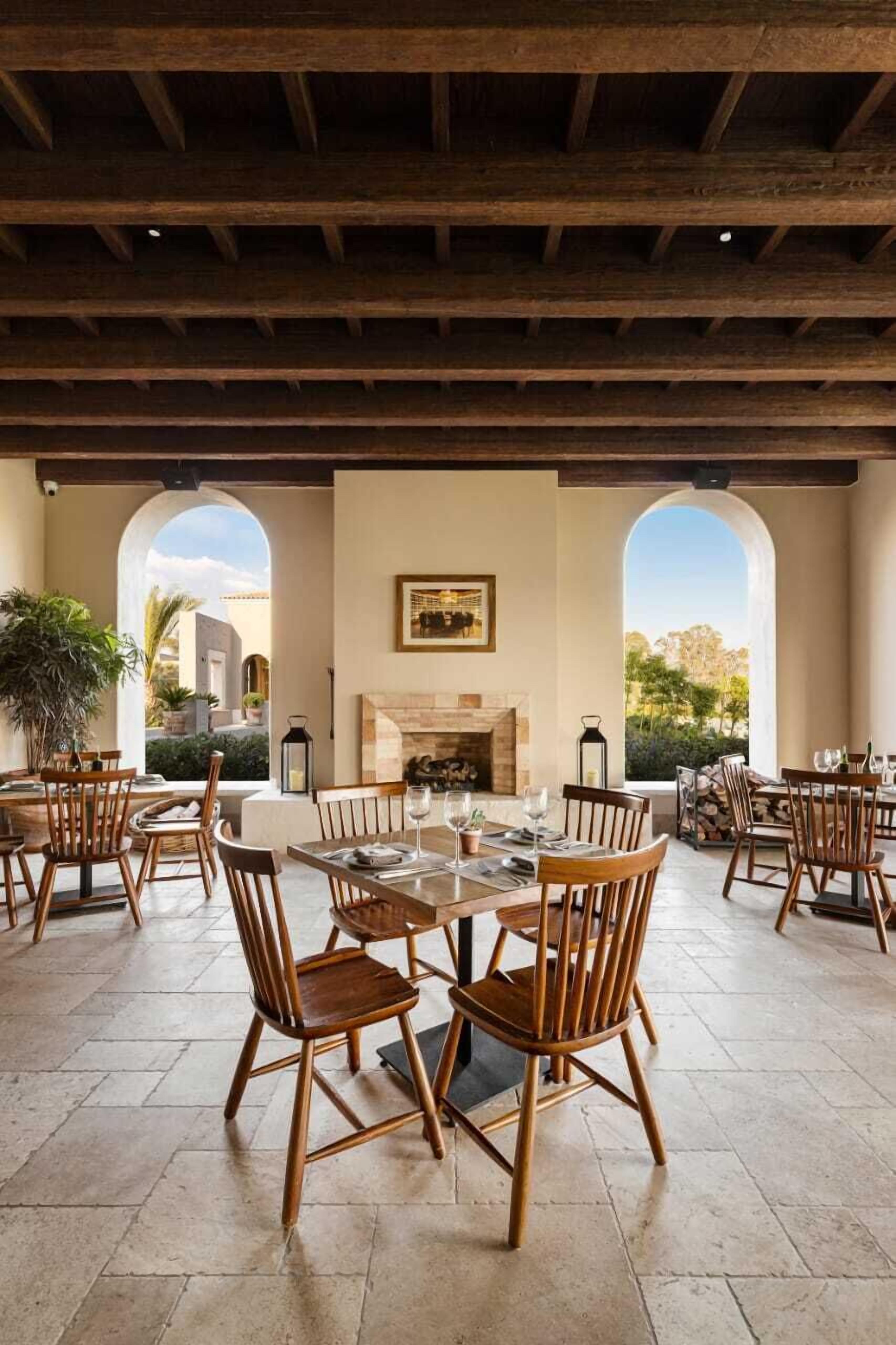An outdoor dining area with wooden chairs and tables, a fireplace, and arched windows overlooking a garden.