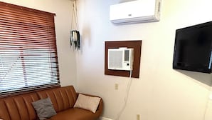 Uma sala de estar com um sofá marrom, uma poltrona rosa, duas mesinhas de cabeceira de madeira, um tapete estampado e uma televisão fixada na parede.