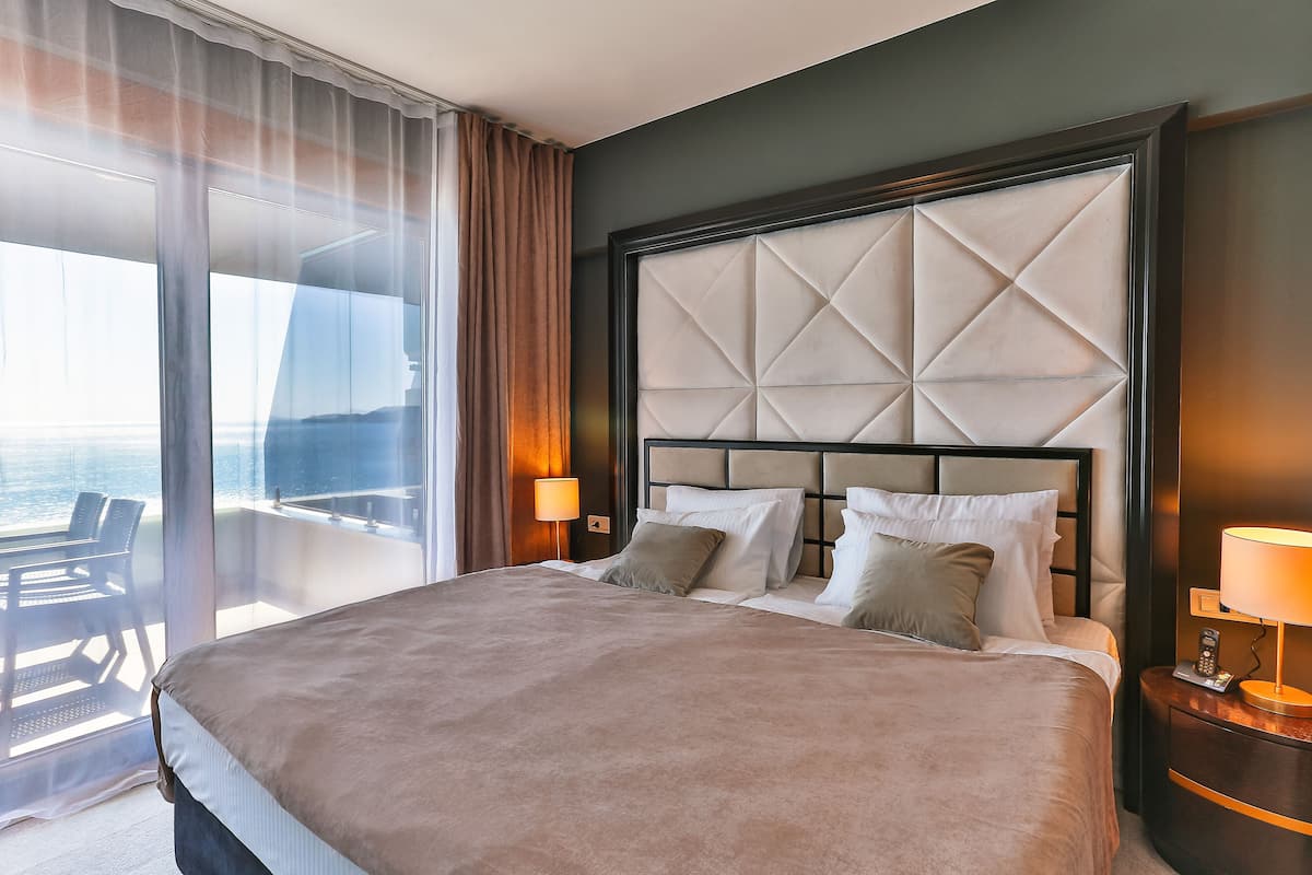 A hotel room with a large bed, a headboard with a geometric pattern, a bedside table with a lamp, and a view of the sea through a large window.