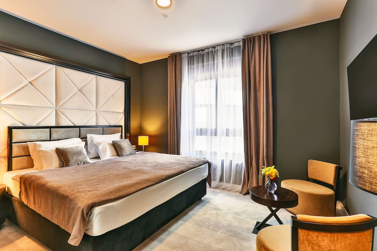 A hotel room with a large bed, a bedside table with a lamp, a chair, and a large window with curtains.