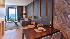 Una habitación de hotel moderna con una mesa de comedor de madera, un cuenco de fruta, una botella de champán y vistas al océano a través de un ventanal grande.