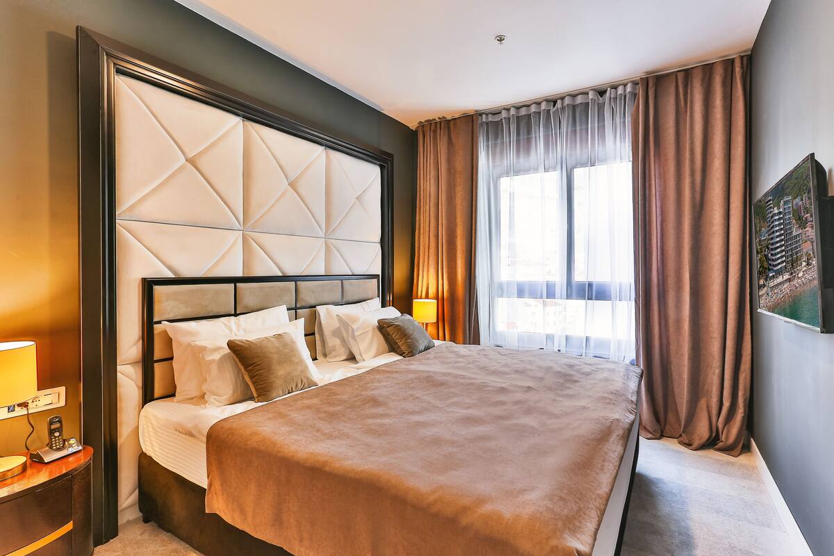 A hotel room with a large bed, a headboard with a geometric pattern, a bedside table with a lamp, and a window with curtains.