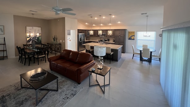 A spacious living room with a brown leather sofa, a dining area with a glass table, and a kitchen with modern appliances and pendant lighting. - Ultra clean designer home in The Villages (3-bed) (Wildwood)