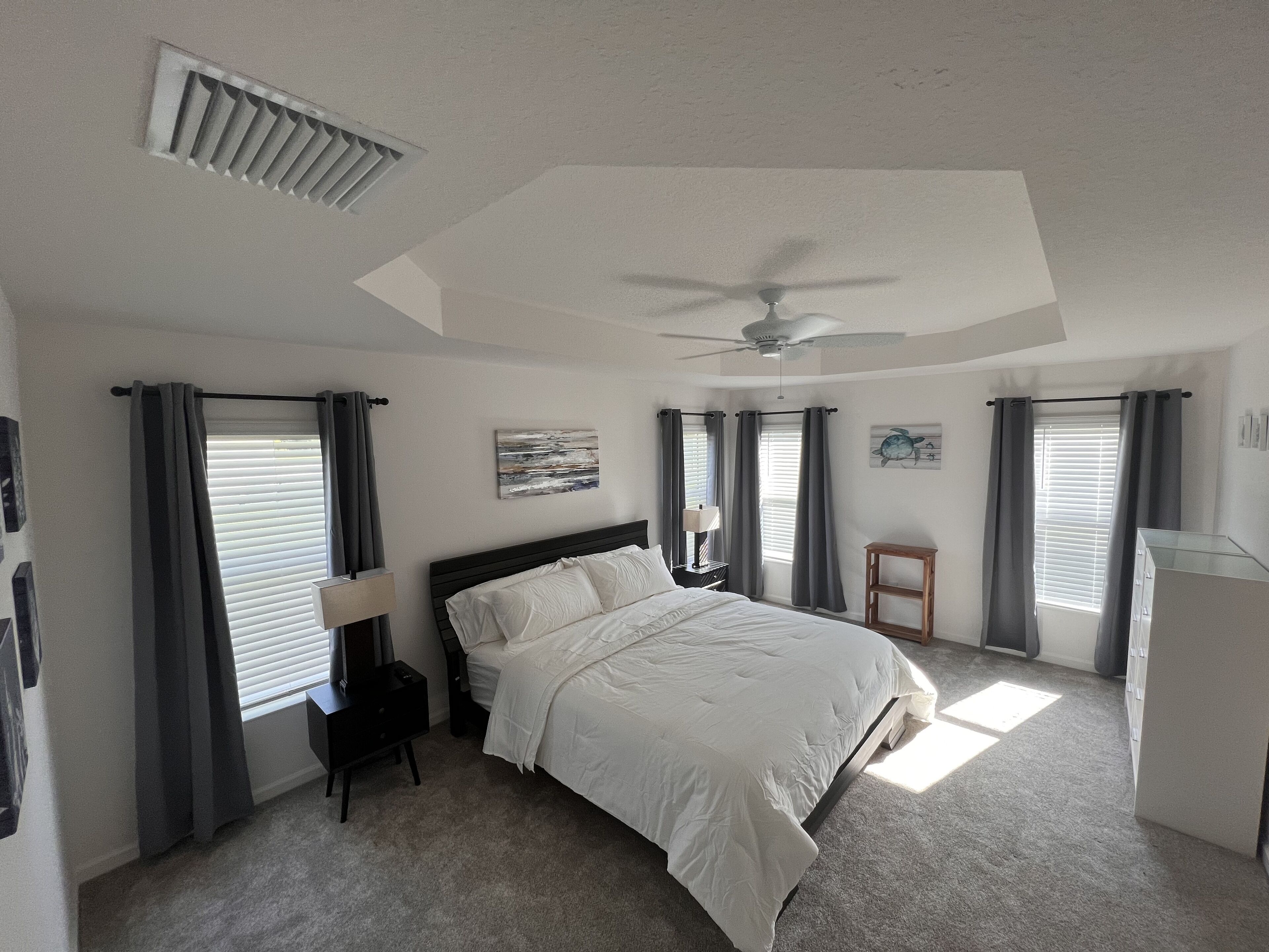 A bedroom with a bed, a nightstand, a lamp, a ceiling fan, and two windows with curtains.