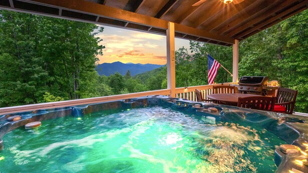 Un bain à remous avec vue sur les montagnes et les arbres, un drapeau américain et une terrasse en bois avec une table et des chaises.