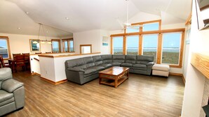 A spacious living room with a large L-shaped gray sectional sofa, a wooden coffee table, and a dining area with wooden chairs and a small island. - Oceanfront, Private Pool, Elevator, Hot Tub, Ocean Views! (Waves)