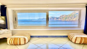 Ein Zimmer mit großem Fenster und Blick auf die Küste, mit Fliesenboden und zwei gestreiften Ottomanen.