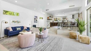 Un salón amplio con un sofá azul, sillones rosados y una mesa de centro. Se observa una cocina al fondo con una zona de comedor.