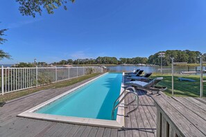 Ein rechteckiger Swimmingpool mit Holzterrasse, umgeben von einem weißen Zaun, mit Blick auf ein Gewässer und Bäume im Hintergrund.