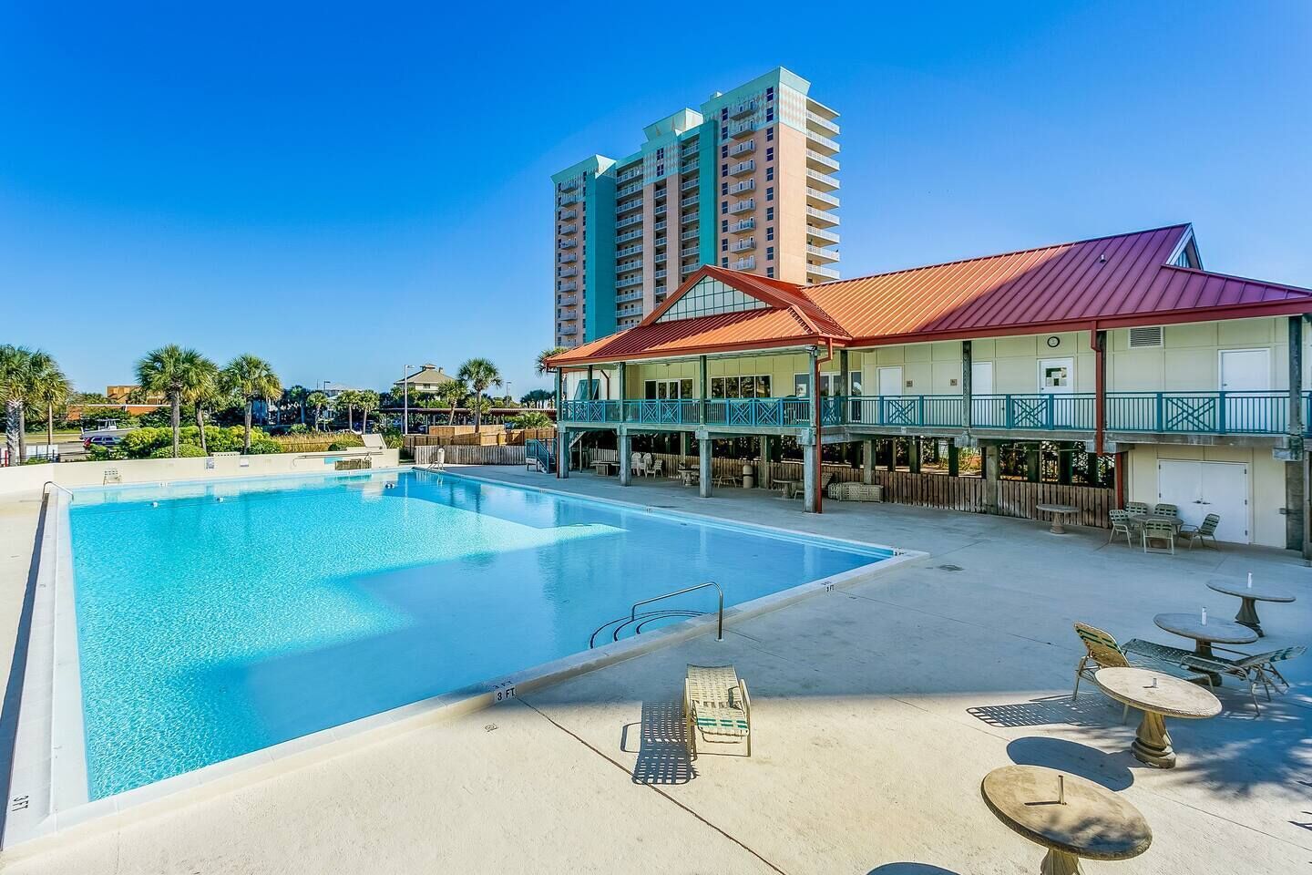 A swimming pool with lounge chairs and tables, a building with a red roof, and a tall residential building in the background.