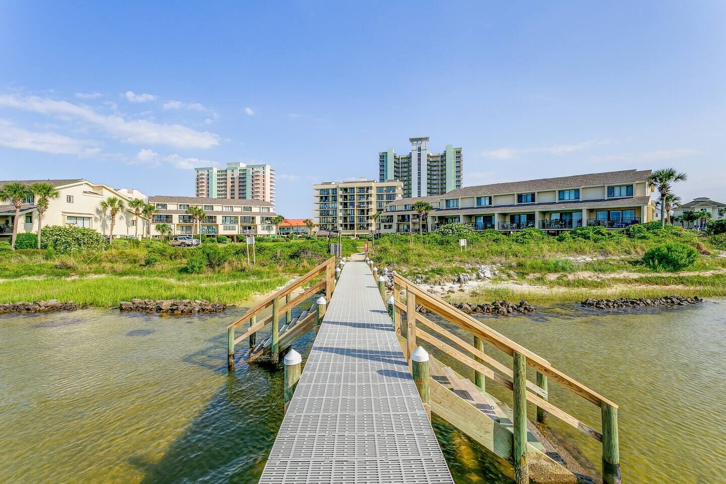 Un puente de madera que conduce a un hotel frente al mar, con varios edificios y una vista despejada del océano.