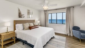 A bedroom with a large bed, a nightstand, a chair, a ceiling fan, and a window with a view of the sea.