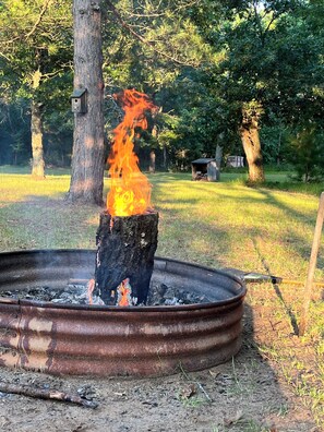 Un feu de camp dans un foyer métallique, entouré d’arbres et d’herbe.