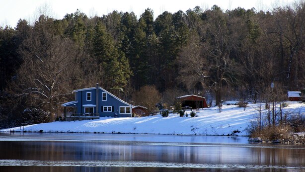 Ein Haus am See mit verschneiter Landschaft und Bäumen im Hintergrund.