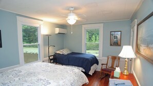 Ein Schlafzimmer mit zwei Betten, einem Deckenventilator, einem Nachttisch mit Lampe und einem Fenster mit Blick auf Grünflächen.