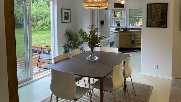 A modern kitchen with a large island, a dining area with a round table and chairs, and a view of a lush garden through the sliding glass doors. - Spacious house in serene peaceful setting 15 min from Lake Crescent&Crescent Bch (Port Angeles)