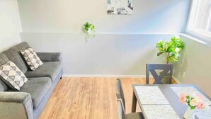 Een woonkamer met een grijze bank, een houten vloer, een eettafel met stoelen en een kleine plant op de vensterbank.