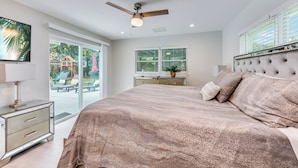 A bedroom with a large bed, a ceiling fan, a TV, and a view of the outdoors.