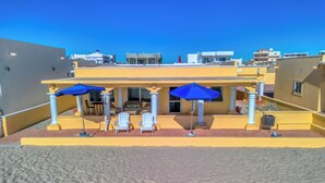 Hotel met een overdekt buitengebied met witte stoelen en blauwe parasols, een zandstrand op de voorgrond en een helderblauwe lucht.
