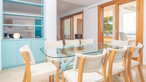 A modern dining area with a glass table and white chairs, a light blue cabinet, and a view of the outdoors through a glass door. - ED. BELLVEURE 2º A - Beautiful apartment on the seafront. Free WiFi. (Pollença)