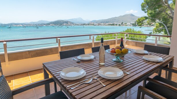 A wooden table set for dining with plates, cutlery, and a bottle on a balcony overlooking a coastal view with boats and mountains. - ED. BELLVEURE 2º A - Beautiful apartment on the seafront. Free WiFi. (Pollença)