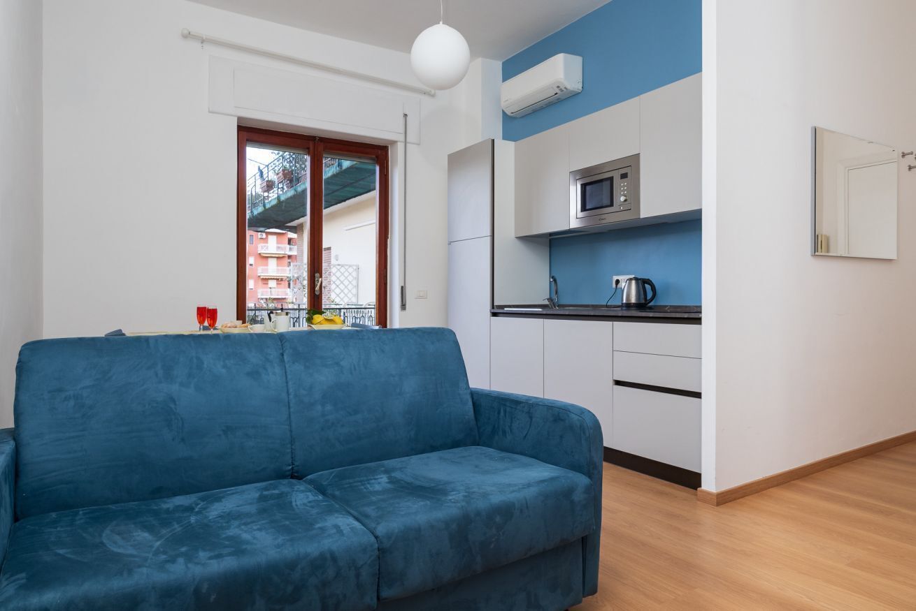 Een moderne woonkamer met een blauwe bank, een keuken met witte kasten en een raam met uitzicht op gebouwen.