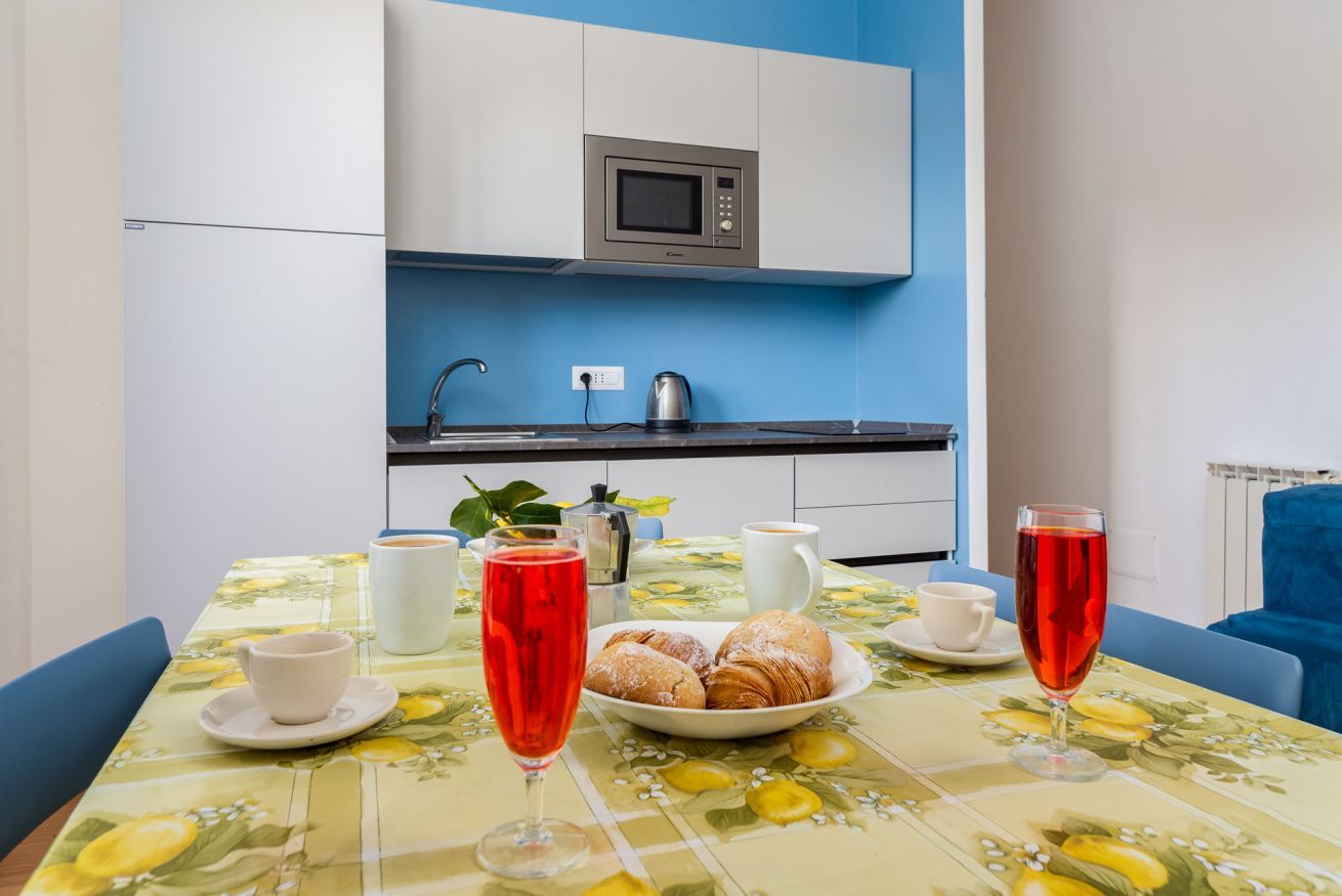 Une table à manger recouverte d’une nappe à motifs floraux, une assiette de pâtisseries, deux verres contenant une boisson rouge, une tasse blanche et une bouilloire.