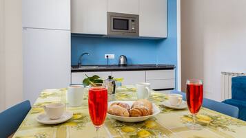 Une table à manger recouverte d’une nappe à motifs floraux, une assiette de pâtisseries, deux verres contenant une boisson rouge, une tasse blanche et une bouilloire.