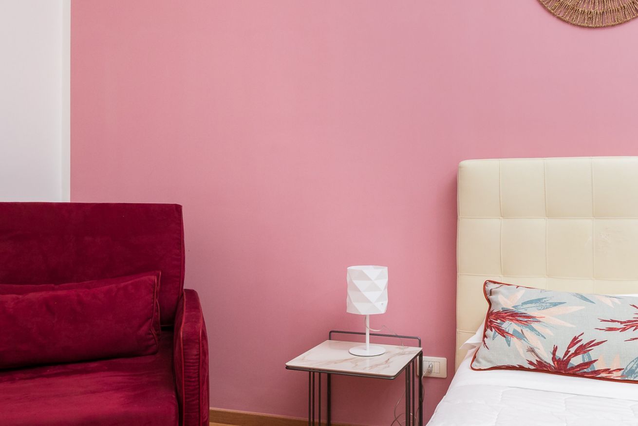 Ein Zimmer mit einem roten Sessel, einem kleinen Tisch mit einer weißen Lampe, einem Bett mit einem Blumenkissen und einer rosa Wand.