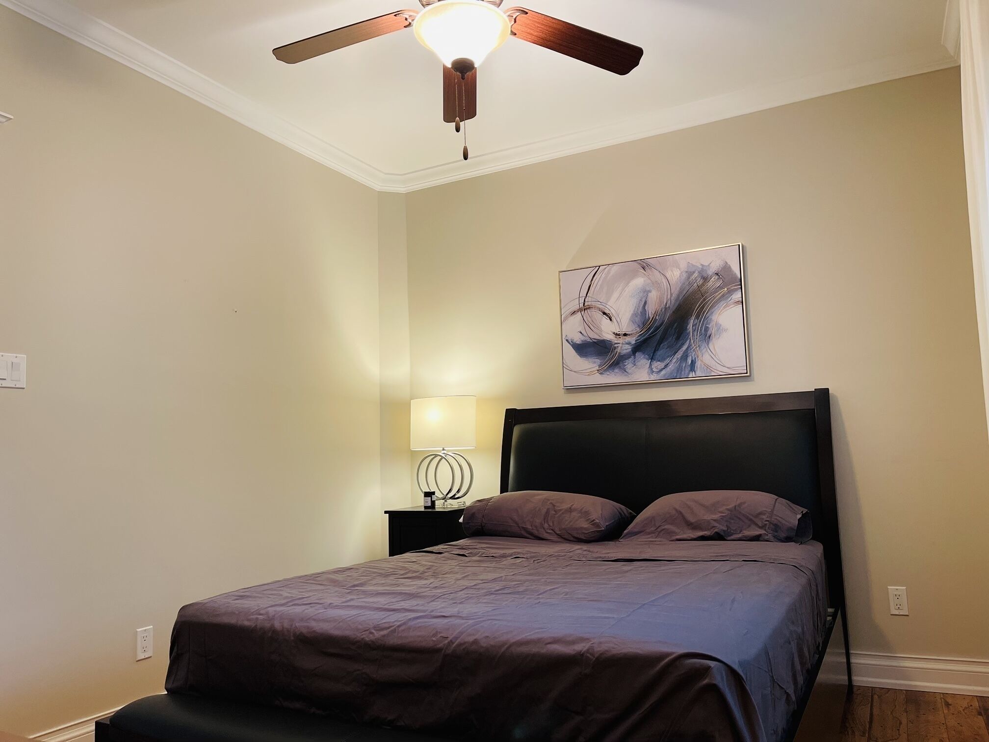 A bedroom with a bed, a ceiling fan, a wall-mounted lamp, and a painting.