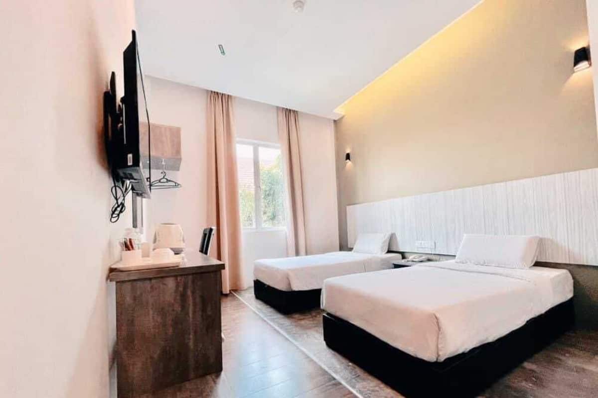 A hotel room with two beds, a desk with a chair, a flat-screen TV mounted on the wall, and a window with curtains.