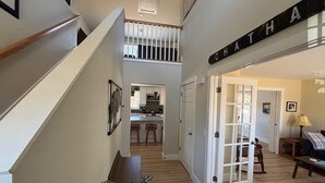 Amplia sala de estar con una escalera que conduce a un segundo piso, una cocina al fondo y una puerta con un letrero que dice "CHATHA".