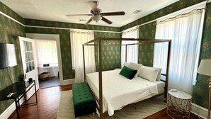 Una cama con dosel, un banco tapizado en verde, una mesita redonda y un ventilador de techo en una habitación con papel tapiz verde y suelo de madera.